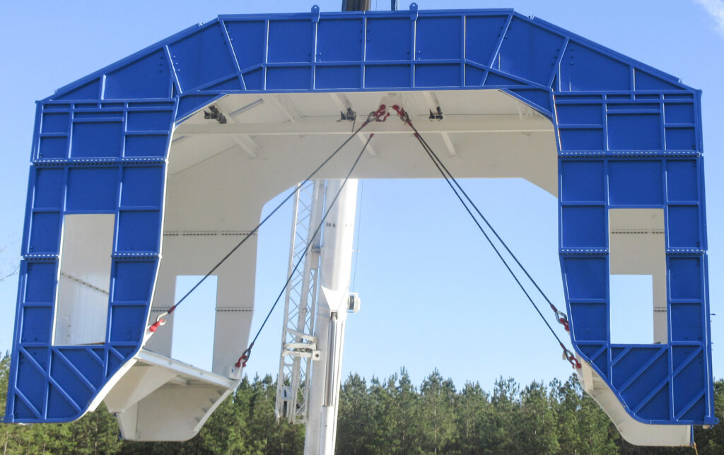 A large blue metal structure with cables attached, suspended outdoors against a clear sky and trees in the background.